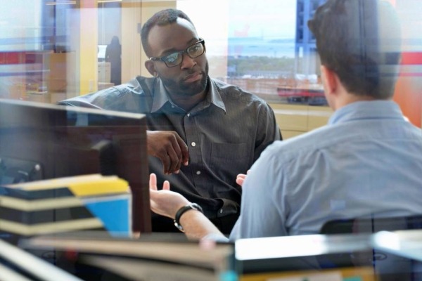  Two colleagues sitting in an office setting and talking with each other.