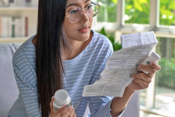 Woman sitting in living room and reading medical instructions in one hand and holding a prescription bottle in the other.